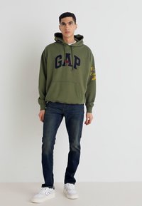 Olivgrön hoodie med "GAP"-logga i marinblått, känguruficka, dragsko i huvan; kombinerad med mörkblå denimjeans och vita sneakers.