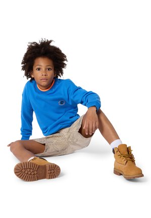 Enfant aux cheveux bouclés assis par terre, portant un sweat-shirt bleu, un short beige à motifs, des chaussettes blanches et des bottes à lacets couleur sable.
