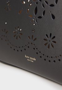 Black leather handbag featuring laser-cut floral designs with small perforations. Gold embossed brand logo at the bottom.