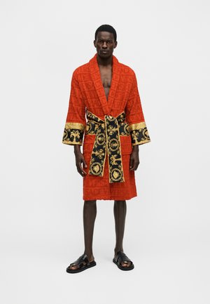 Άνδρας που φορά κόκκινο μανδύα με μαύρη και χρυσή διακοσμημένη τρέσα και ζώνη, στέκεται ξυπόλητος σε απλό λευκό φόντο.