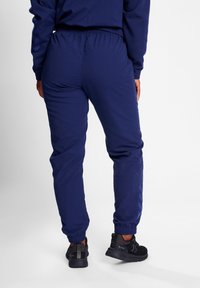 Marina blå idrottsbyxor med elastisk midja, sidostrimmor och avslutade anklar, kombinerade med svarta idrottsskor.