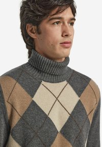 Grauer Rollkragenpullover mit einer strukturierten Rippoptik, der ein Rautenmuster in Beige- und Brauntönen aufweist, was einen Kontrast in Farbe und Design hervorhebt.