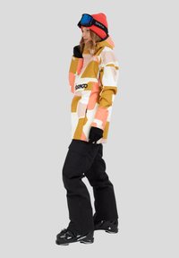 Personne portant une veste de ski à motifs orange et beige, un pantalon de neige noir, des bottes de ski noires, un bonnet rouge et des lunettes de ski sur la tête.