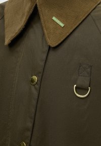 Gros plan sur une veste olive foncé avec un col en velours marron, une épingle verte étiquetée SPEY, des boutons en laiton et un détail en anneau en D sur la poitrine.