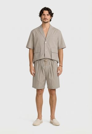 Uomo in piedi con camicia beige a maniche corte abbottonata, pantaloncini con coulisse coordinati e scarpe leggere slip-on su sfondo semplice.