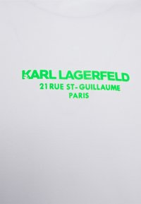 Camiseta de algodón blanca con texto verde en negrita: "KARL LAGERFELD 21 RUE ST-GUILLAUME PARÍS." Textura suave con un diseño moderno.