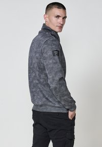 Hombre con chaqueta gris de diseño a rayas y cremallera, con alto cuello y pantalones cargo negros, mirando por encima del hombro contra un fondo claro y liso.