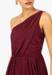 Robe bordeaux à une épaule en tissu doux, présentant une texture drapée et des détails froncés à la taille, créant une silhouette fluide.