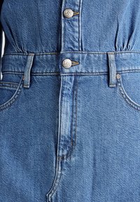 Macacão de denim em azul médio com botões prateados apresentando a marca "Lee". Inclui bolsos laterais e um elástico na cintura definido para estrutura.