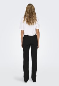 Zwarte op maat gemaakte broek met een gladde textuur, met een slanke pasvorm en gestructureerd ontwerp. G gedragen met een losse witte T-shirt, gezien van achteren.