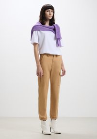 T-shirt bianca a maniche corte, pantaloni cargo color cammello con tasche laterali, e un maglione lilla chiaro appeso sulle spalle. Stivali bianchi con tacco alto.