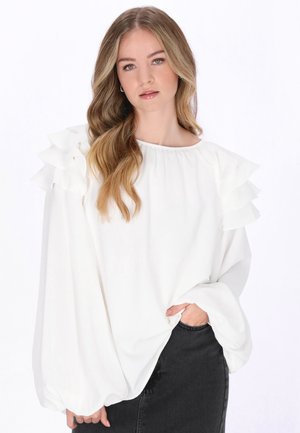 Młoda kobieta o długich, falowanych blond włosach nosi białą bluzkę z falbankami na ramionach i obszernymi rękawami, do której dobrała czarną spódnicę.