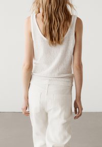 Top in cotone bianco a costine con schiena a scollo, indossato con pantaloni bianchi abbinati e texturizzati, che mettono in evidenza linee orizzontali sottili e tasche pratiche.