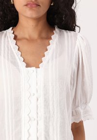 Blouse blanche avec un tissu texturé à rayures verticales, un trim en dentelle festonnée le long de l'encolure et des manches, et des détails de boutons.