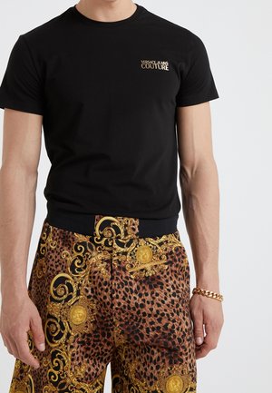 Camiseta negra de algodón con mangas cortas que presenta un pequeño logo dorado. Combinada con pantalones marrones y dorados con estampado de leopardo y detalles barrocos.