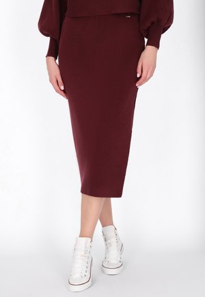 Femme portant une jupe crayon midi bordeaux et un pull oversize assortis à des baskets montantes blanches, debout, jambes croisées.