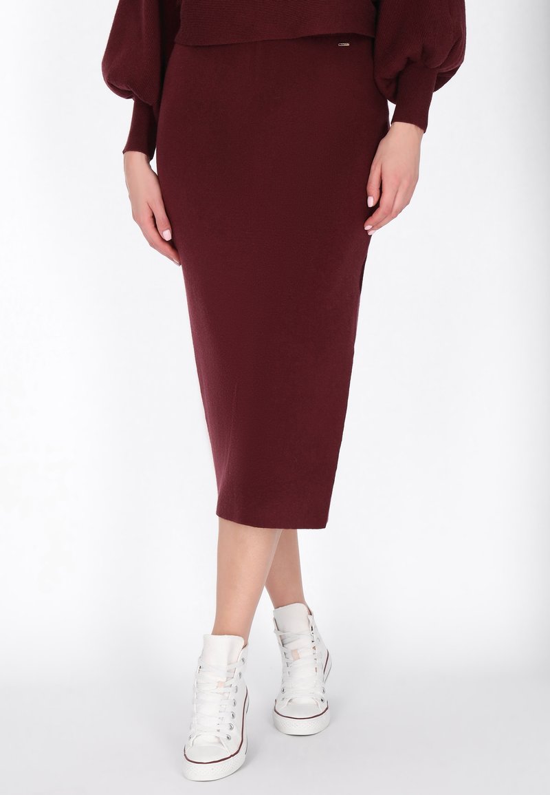 Femme portant une jupe crayon midi bordeaux et un pull oversize assortis à des baskets montantes blanches, debout, jambes croisées.