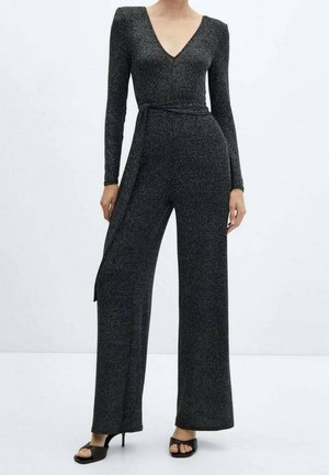 Kobieta ubrana w czarny, błyszczący kombinezon z długim rękawem, głębokim dekoltem w serek i szerokimi nogawkami, do tego czarne sandały na obcasie z odkrytymi palcami.