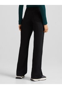 Pantalones de pierna ancha negros, hechos de una tela suave, con bolsillos traseros y una cintura alta. El modelo lleva zapatillas con suela blanca.