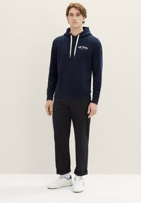Felpa blu navy con cappuccio e coulisse, con logo sul petto, abbinata a pantaloni neri e sneakers bianche. Tessuto liscio, vestibilità casual, senza motivi visibili.