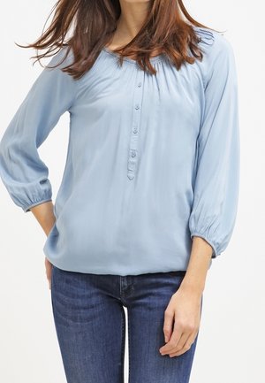 Femme portant un chemisier bleu clair à manches longues avec des boutons et un jean bleu foncé, debout avec une main sur la hanche.