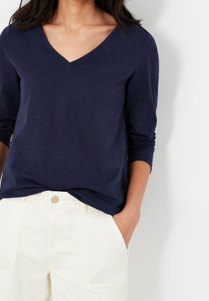 Kobieta ubrana w granatową koszulę z długim rękawem i dekoltem w serek oraz kremowe spodnie, z jedną ręką w kieszeni, na tle jednolitego jasnego tła.