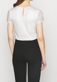 Top blanco de encaje con mangas cortas, que presenta un detalle de abertura en la parte posterior, combinado con pantalones negros ajustados. Patrón floral texturizado visible.