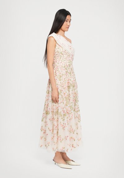 Vrouw in een bloemrijke, een-schouder maxi-jurk met plooien, gekleed in crèmekleurige puntige slingback hakken, tegen een eenvoudige witte achtergrond.