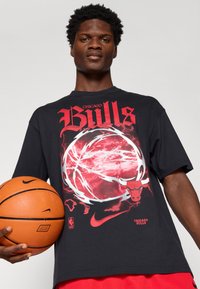 Camiseta preta de manga curta com um gráfico vermelho de uma bola de basquetebol com chamas e o texto "Chicago Bulls". A segurar uma bola de basquetebol.
