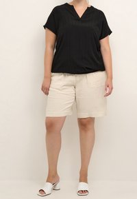 Haut noir à manches courtes avec des rayures verticales, décolleté en V ; associé à un short en lin beige avec une poche latérale. Sandales à talons blanches à enfiler.