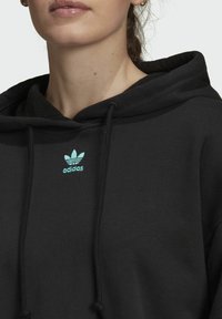 Svart bomullshuvtröja med känguruficka, snören och en ljusgrön broderad Adidas-logga på bröstet. Mjuk textur.