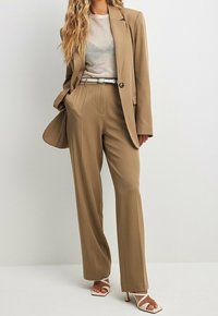 Bronzový oblek na míru s blejzrem a kalhotami s širokými nohavicemi, doplněný světle šedou košilí a bílými sandály na podpatku s pásky, s hladkou texturou.