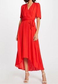 Rode wikkeljurk met korte mouwen, een strik taille en een asymmetrische zoom. Gladde stof met een subtiele glans, gecombineerd met sandaaltjes met bandjes.