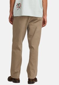 Pantalons en coton beige avec une coupe droite et des coutures visibles. Portés avec des chaussures noires et beiges à motifs. Détail brodé au bord de la chemise.
