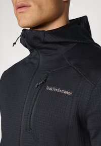 Giacca nera con cappuccio realizzata in tessuto testurizzato, tasca con zip e branding discreto sul petto. Presenta un cordino regolabile al collo.