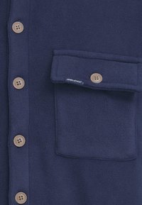 Camisa de forro azul marino con cierre de botones, botones beige y un bolsillo en el pecho con etiqueta. Textura suave y diseño uniforme.