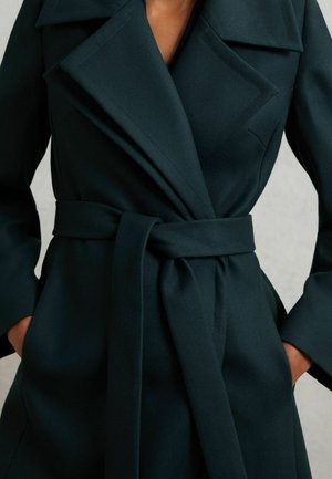 Fekete öves kabát széles hajtókkal, a zsebébe dugott kézzel viselve, kiemelve a testre szabott illeszkedést és a sima szövet textúrát.