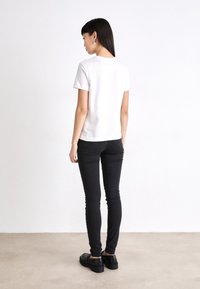 Sieviete ar gariem taisniem matiem, kurai ir sudraba riņķa auskari, baltā īsā kreklā, melnās šaurās biksēs un melnās mokasīnās, stāv ar muguru pret skatītāju.