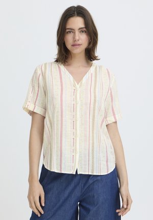 Kvinde med skulderlangt brunt hår iført en kortærmet cremefarvet bluse med lodrette røde og beige striber og blå bukser.