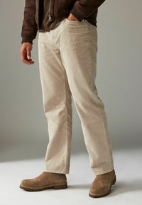 Pantalons en velours côtelé beige avec une coupe droite, présentant une surface texturée et des poches latérales, associés à des bottines marron.