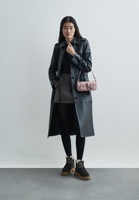 Trench-coat en cuir noir avec un devant boutonné, associé à une jupe marron, des collants noirs et des bottes marron ornées de fourrure. Sac à main rose.