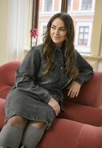 Robe en denim gris foncé avec fermeture à boutons, poches poitrine et ceinture à nouer, portée avec des chaussettes grises montantes sur une chaise en tissu rose.