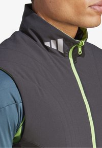 Chaleco sin mangas de tela gris oscuro con cremallera verde, que presenta un alto cuello y un sutil patrón acolchado. Acentos de logo en blanco.