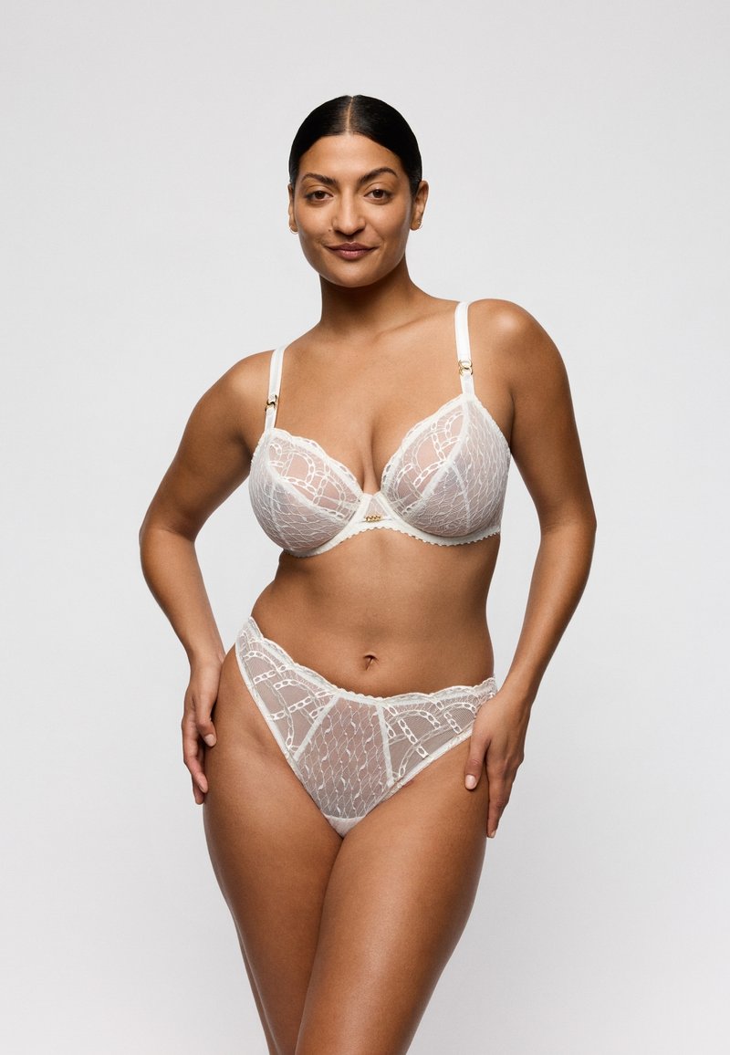 Mujer modelando un sujetador de encaje blanco y ropa interior a juego de talle alto, de pie con las manos en las caderas contra un fondo claro y liso.