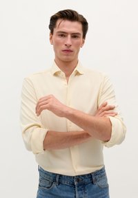 Light yellow, long-sleeve button-up shirt with a soft texture. Features rolled sleeves and a subtle logo, paired with blue jeans.