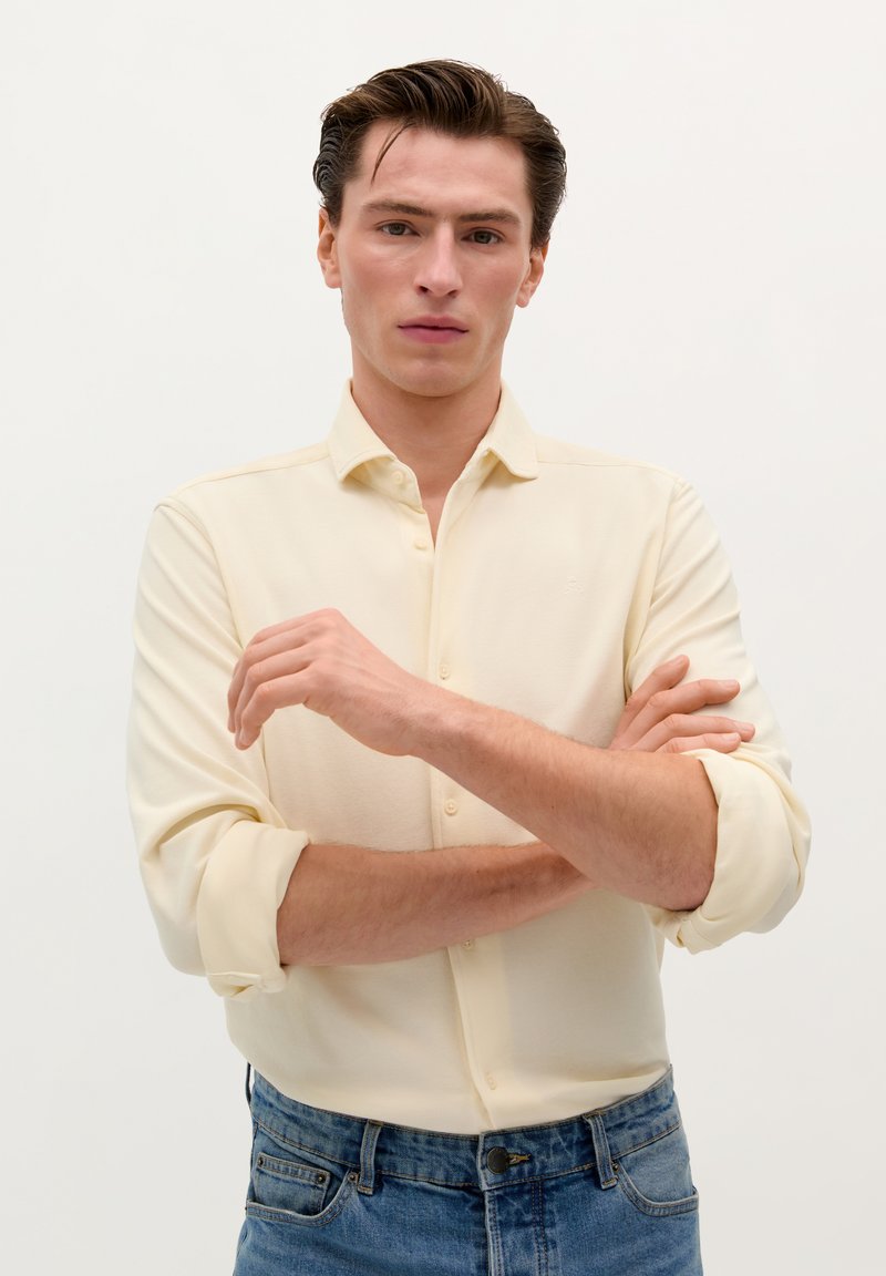 Light yellow, long-sleeve button-up shirt with a soft texture. Features rolled sleeves and a subtle logo, paired with blue jeans.
