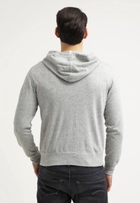 Hombre con sudadera con capucha gris claro y jeans negros, de espaldas contra un fondo blanco liso.