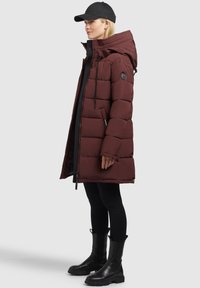 Bordová pérová bunda s kapucňou, ktorá má zips vpredu, bočné vrecká a nášivku s logom. Čierne legíny a masívne čierne čižmy dopĺňajú tento outfit.