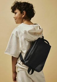 Sac à dos en tissu noir porté sur le dos, avec une fermeture éclair sécurisée et des bretelles réglables. Modèle dans un hoodie léger et ample. Fond neutre.