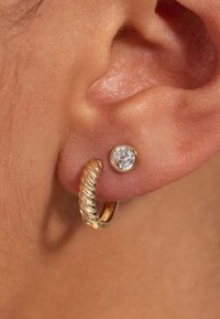 Boucles d'oreilles créoles en ton or avec une texture tordue à côté d'une petite pierre ronde claire sertie dans une puce assortie de ton or.